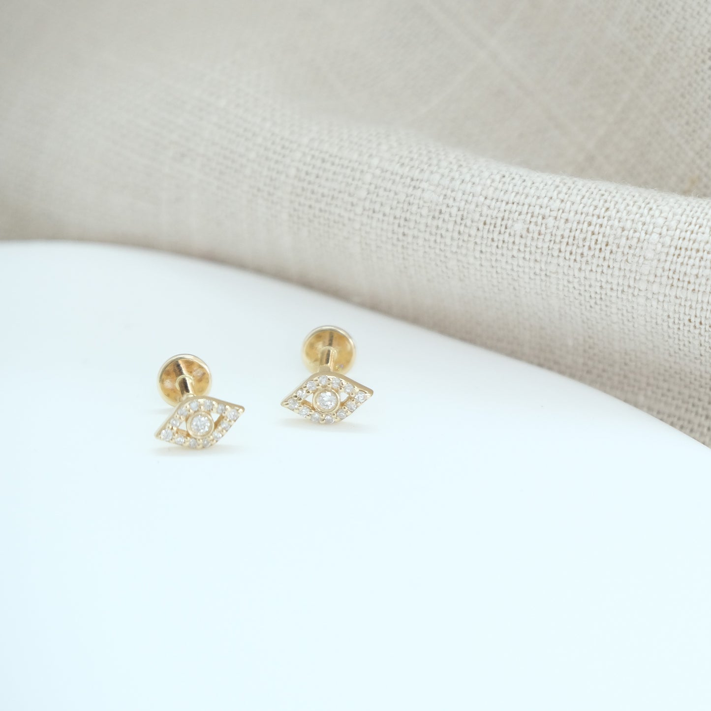 14ct recycled gold diamond evil eye studs on a white stone background and beige linen