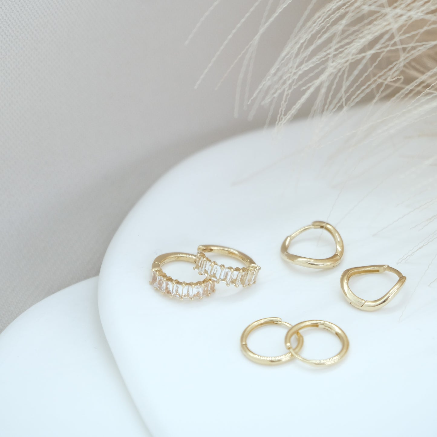 14ct gold classic huggies, wavy huggies and white topaz baguette huggies on a white background with pampas